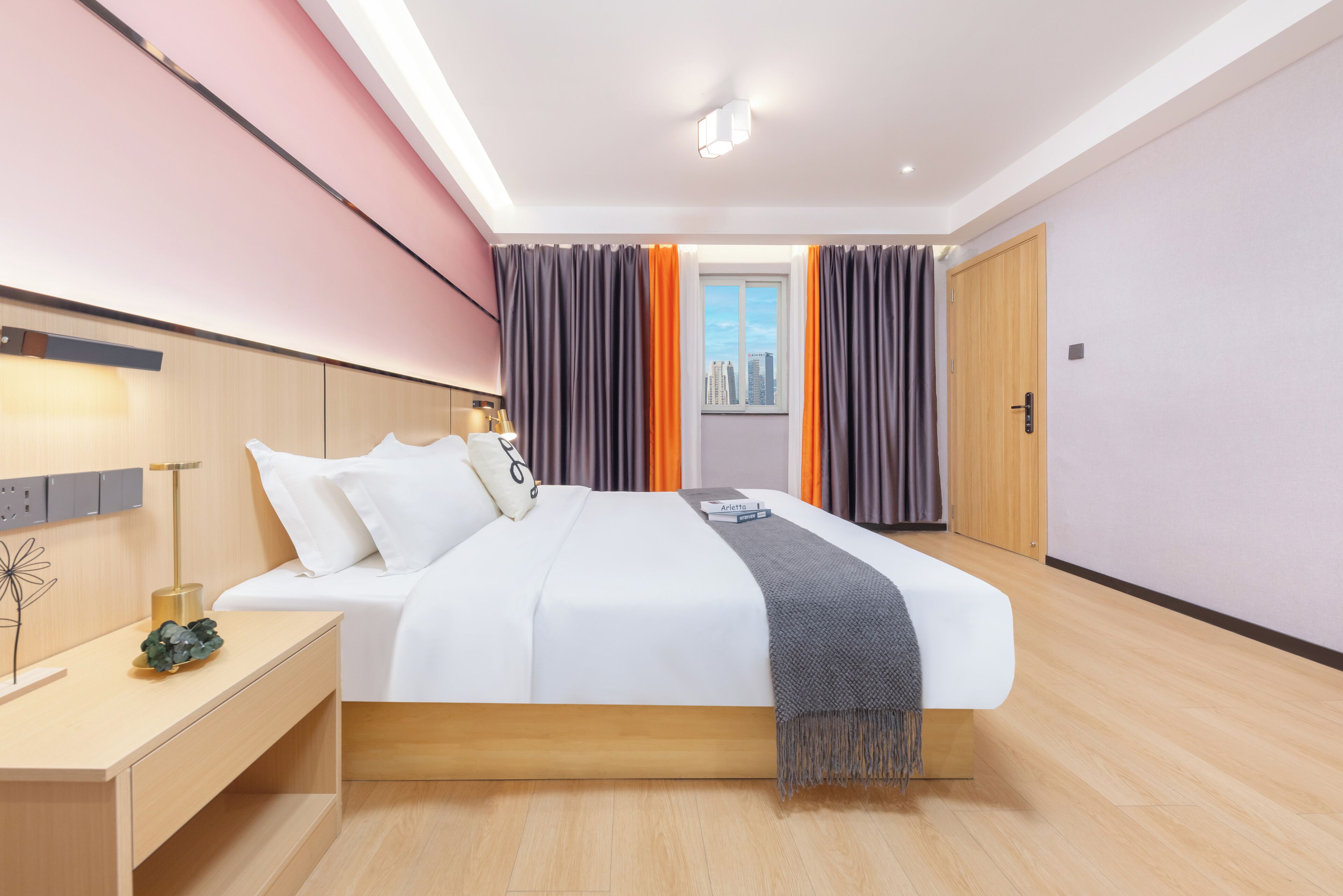 A modern hotel room with a large bed, bedside tables, a nightstand, a city view through the window, and a wooden door.