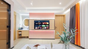 Habitació d'hotel moderna amb televisió de pantalla plana penjada a la paret que mostra diferents canals, una porta de fusta, una taula petita amb objectes decoratius i un finestral gran amb cortines.