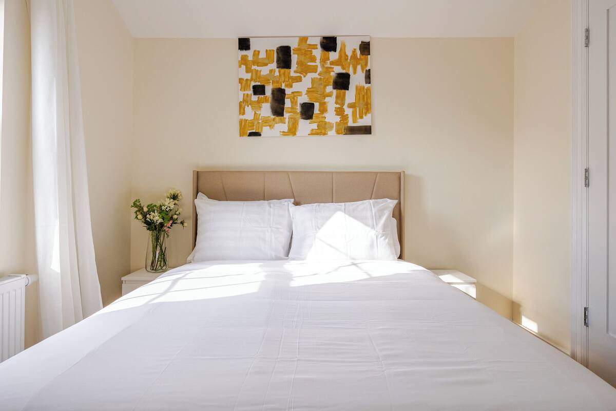 Une chambre d’hôtel avec un grand lit, une tête de lit, une table de chevet ornée d’un vase de fleurs et une œuvre d’art murale.