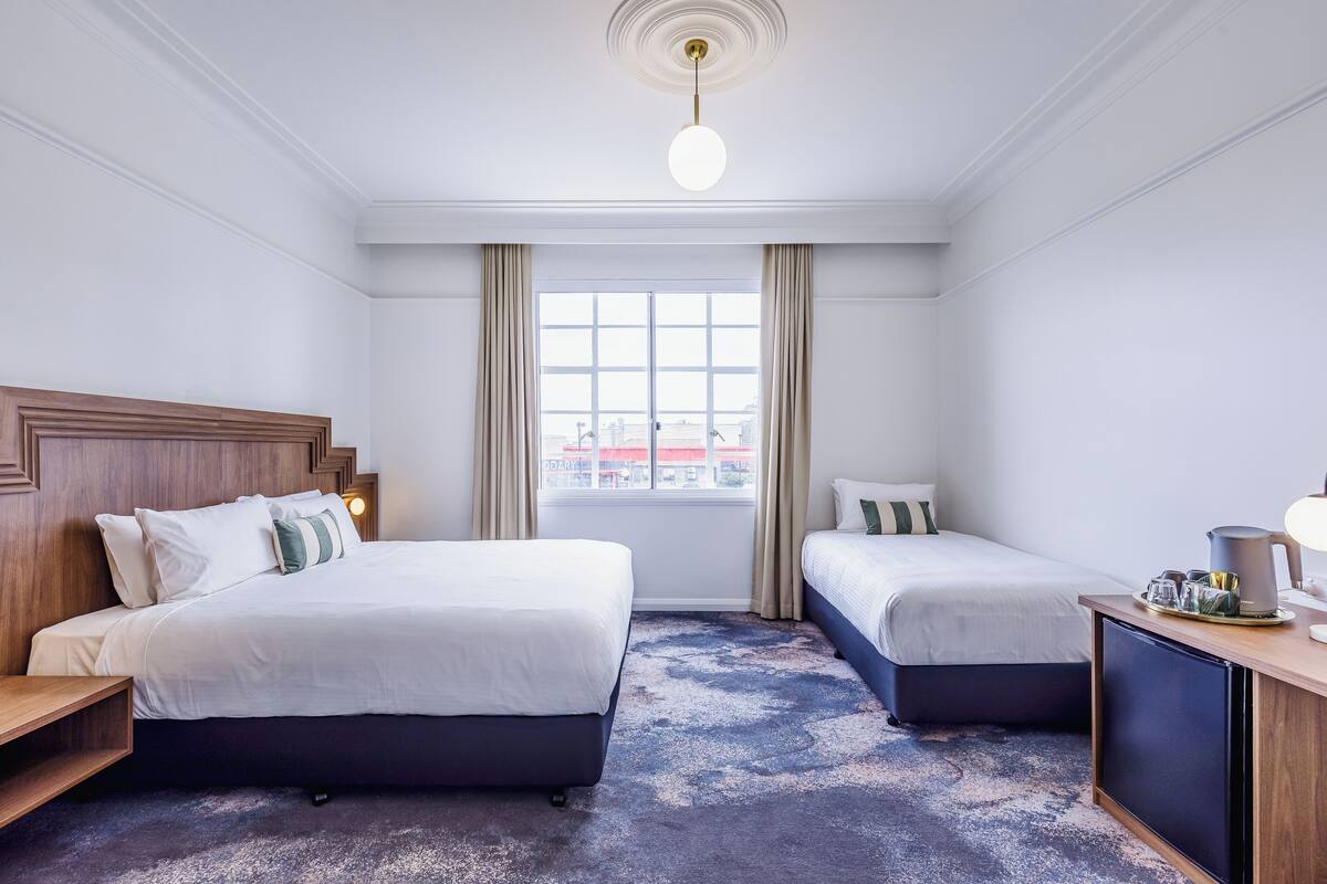 Ein Hotelzimmer mit zwei Betten, einem Holzkopfteil, einem großen Fenster mit Vorhängen, einer Deckenleuchte sowie einem kleinen Schreibtisch mit Lampe und Wasserkocher.