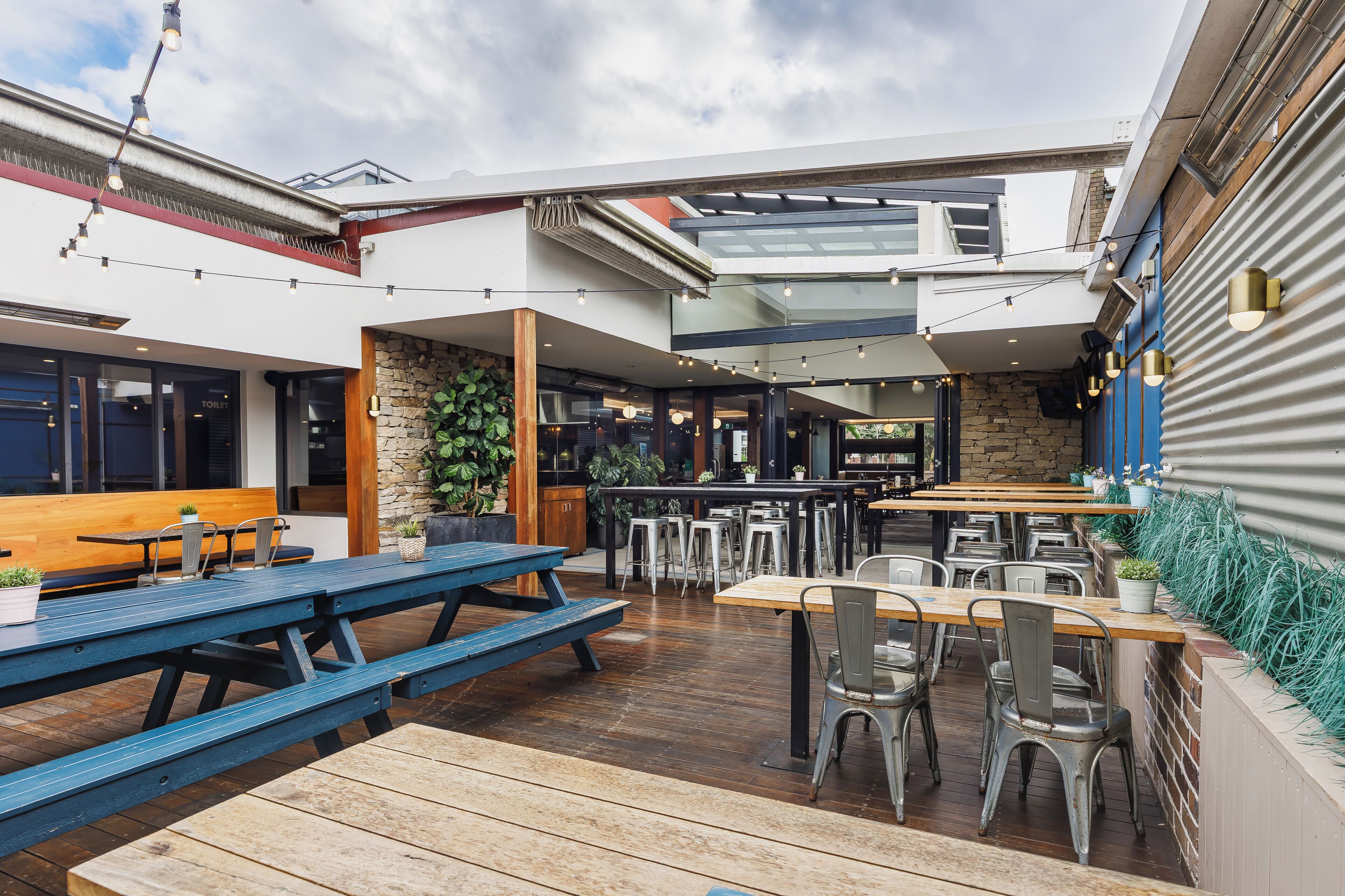 An outdoor patio area with wooden tables and chairs, a stone wall, and a bar area with stools.