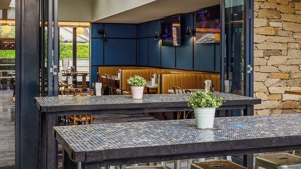 A modern dining area with a high ceiling, exposed beams, and a mix of dark and light-colored walls. There are multiple tables and stools, and a stone wall in the background. - The Lakes Hotel, Rosebery (Rosebery)