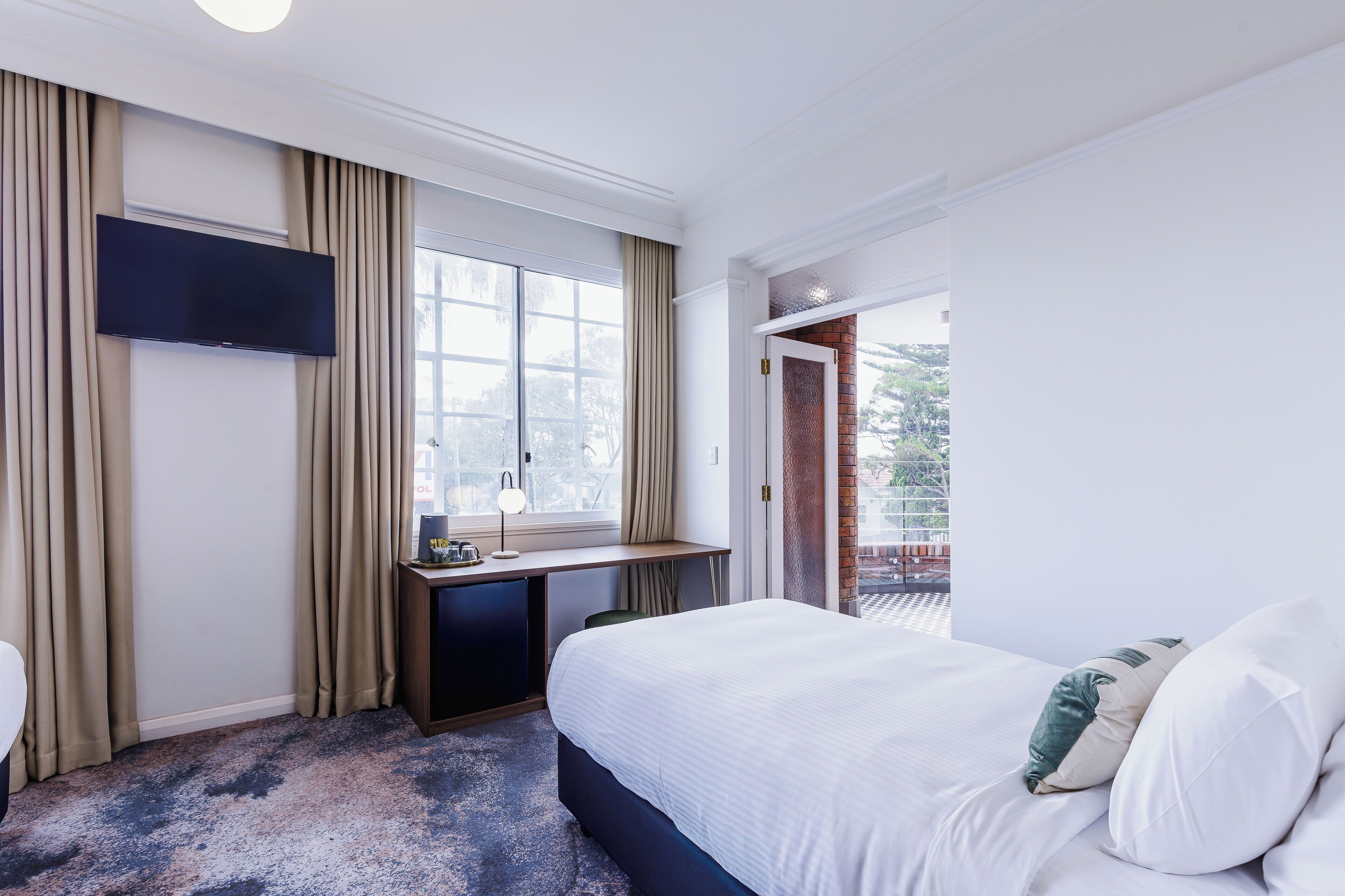 A hotel room with a bed, a desk with a flat-screen TV, a window with curtains, and a view of the outside.