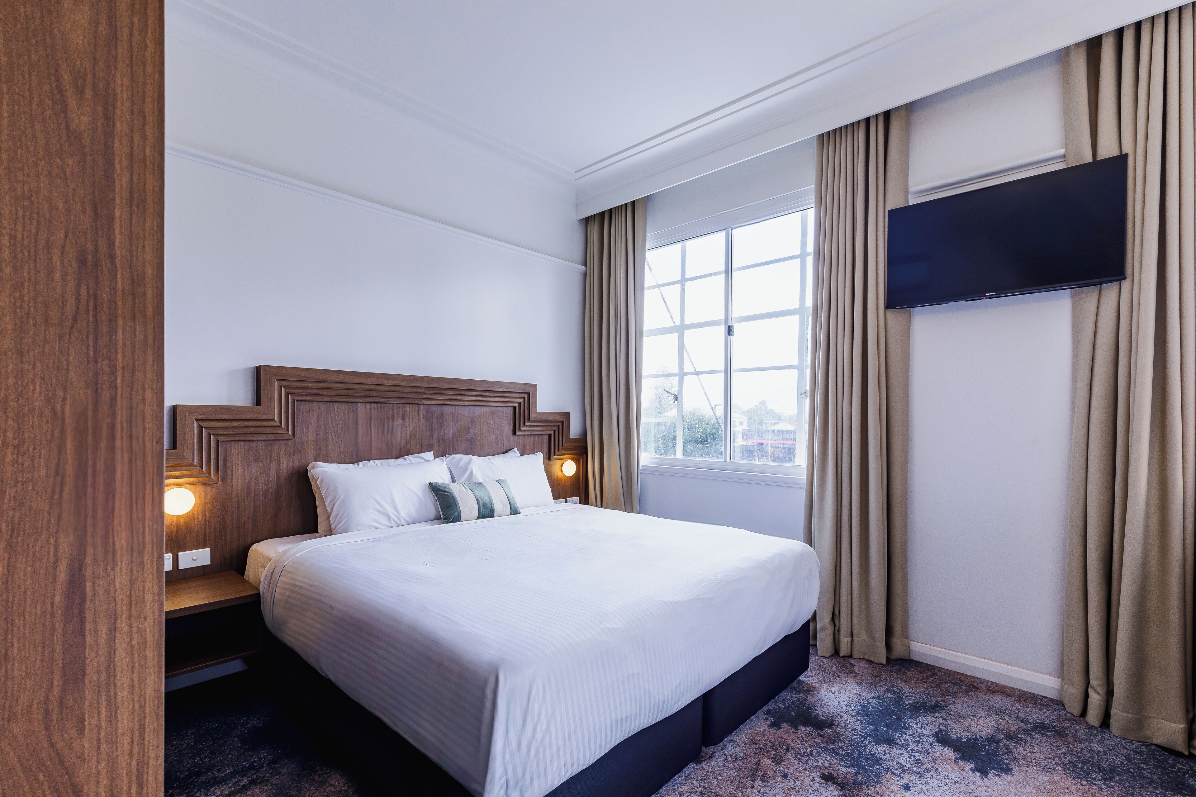 A hotel room with a large bed, a TV mounted on the wall, and a window with curtains.