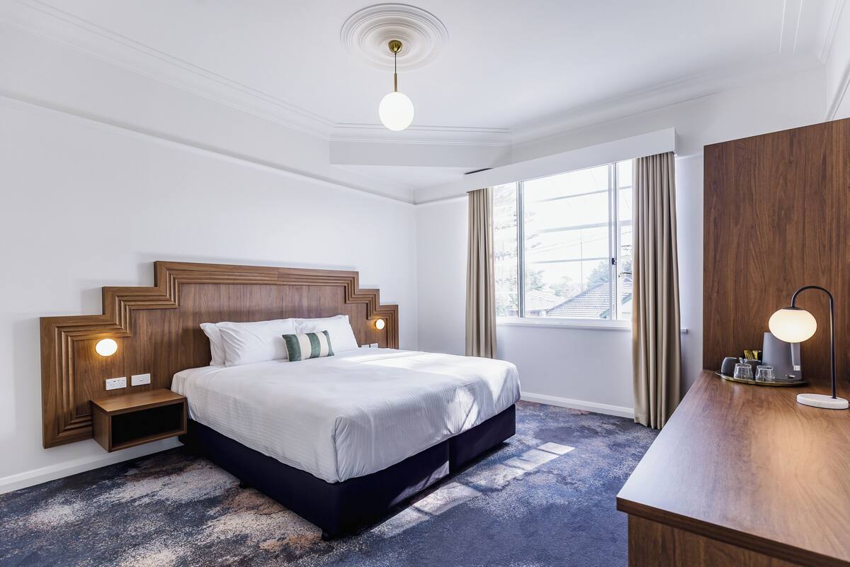 Ein Hotelzimmer mit einem großen Bett, einem Holz-Kopfteil, einem Nachttisch, einem Schreibtisch und einem Fenster mit Vorhängen.