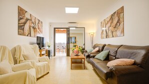 Un salon avec un canapé en cuir marron, un fauteuil blanc et une table basse en bois sur laquelle est posé un vase de fleurs. Deux tableaux ornent les murs et un téléviseur est installé sur un meuble.