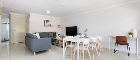 A modern living room with a grey sofa, a white dining table with chairs, a TV, and a small side table with a vase.
