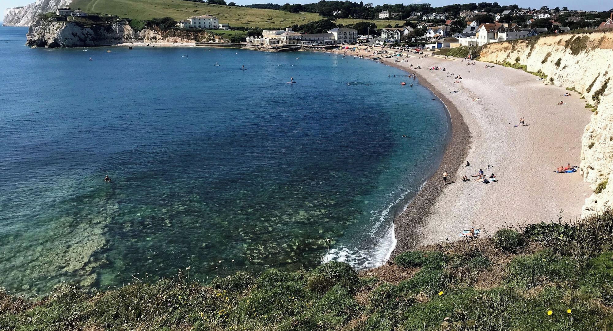 A coastal beach with clear water, a sandy shore, and buildings in the background.