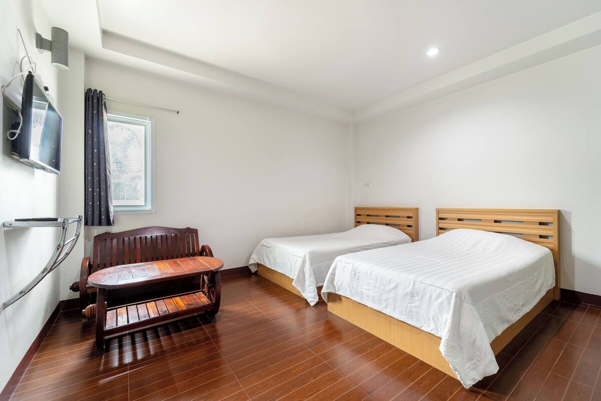 Une chambre d’hôtel avec deux lits simples, un banc en bois, un téléviseur fixé au mur et une fenêtre avec des rideaux.
