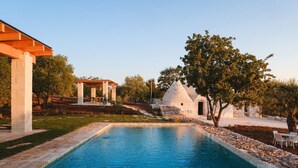 A swimming pool with a stone border, a white building with a conical roof, and a pergola structure. - Luxury Apulian Villa inside Trulli with unique pool (Castellana Grotte)