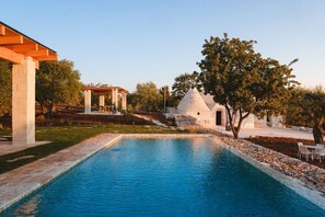 A swimming pool with a stone border, a white building with a conical roof, and a pergola structure. - Luxury Apulian Villa inside Trulli with unique pool (Castellana Grotte)