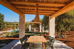 Un patio al aire libre con mesa de madera y sillas de mimbre, un pilar de piedra y vistas a una piscina y jardín.