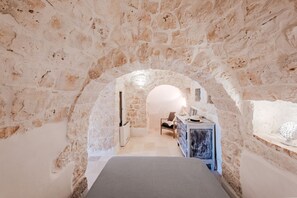A room with a stone wall archway leading to a bed and a desk area.