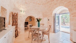 A dining area with a round table, wicker chairs, and a vase of flowers. There is a television on a cabinet to the left. - Luxury Apulian Villa inside Trulli with unique pool (Castellana Grotte)