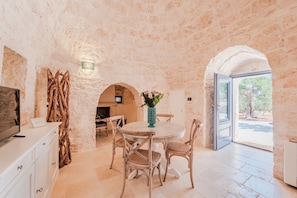 A dining area with a round table, wicker chairs, and a vase of flowers. There is a television on a cabinet to the left. - Luxury Apulian Villa inside Trulli with unique pool (Castellana Grotte)