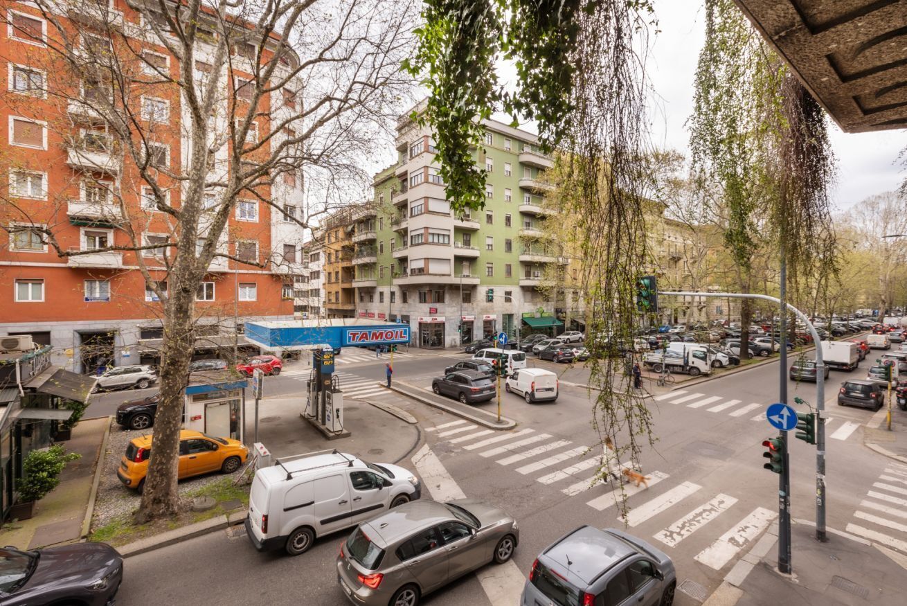 A multi-story building with a green and white facade, a visible storefront with a blue sign, and a street intersection with cars and a pedestrian crossing.