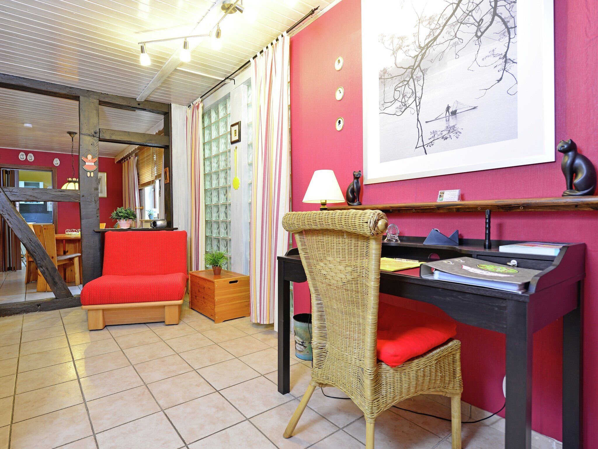 Une pièce chaleureuse avec un canapé rouge, un fauteuil en osier, un bureau avec une lampe et une carte encadrée accrochée au mur.