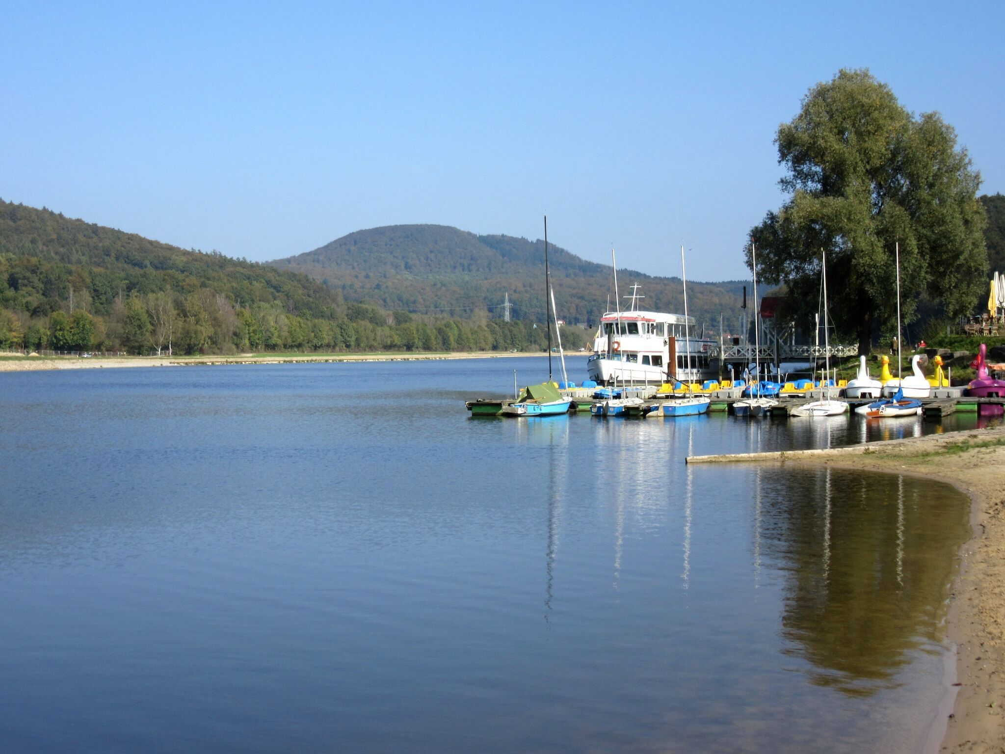 Ein ruhiger See mit mehreren Booten, die am Ufer liegen, umgeben von Hügeln und Bäumen.