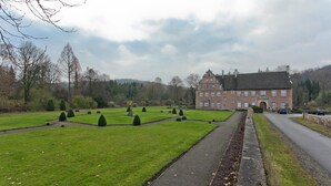 Een groot, historisch gebouw met een symmetrische tuinaanleg, waaronder netjes gesnoeide hagen en een centrale fontein.
