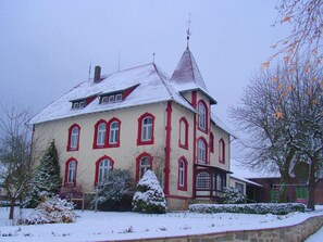 A large, two-story building with a steeply pitched roof and a prominent tower. - Ferienwohnung mit Garten auf dem Bauernhof (Trendelburg-Friedrichsfeld)