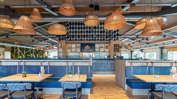 Un ristorante con tavoli in legno e banquette imbottite blu, lampade appese in vimini e una parete decorata.