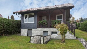 Een woonhuis met twee verdiepingen, een stenen gevel, een houten veranda en een kleine tuin.