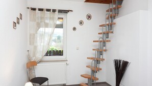 Een kamer met een houten trap, een raam met uitzicht, een stoel, een lamp en een radiator.
