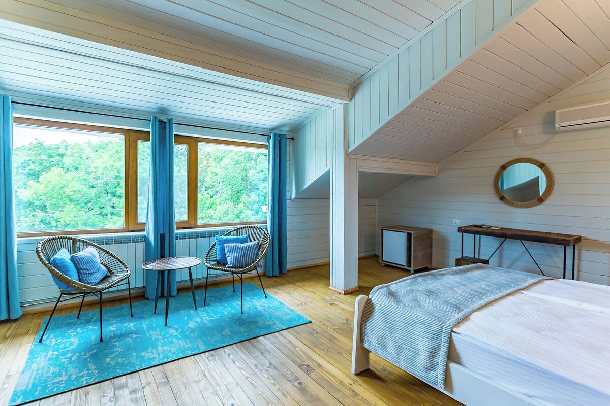 A bedroom with a bed, a small table, two chairs, a mirror, and a window with a view of trees.