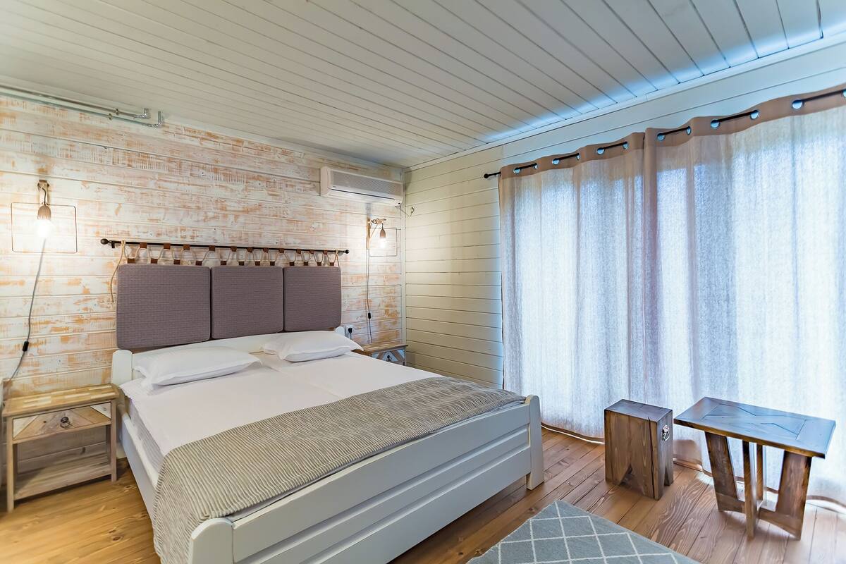 A modern bedroom with a wooden headboard, a bed with white linens, a nightstand, a small wooden table, and a window with curtains.