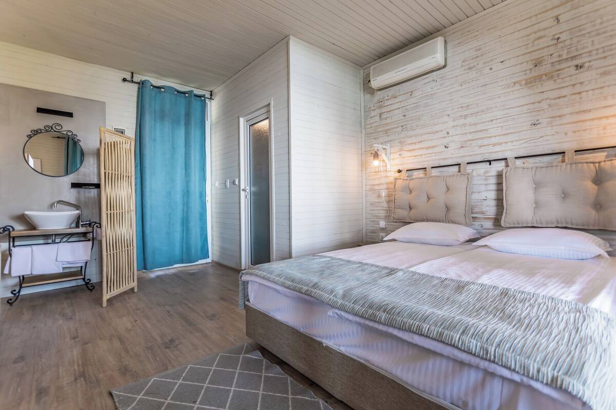 A bedroom with a bed, a bedside table, a mirror, a window with curtains, and a wall-mounted air conditioner.