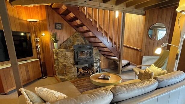 A cozy living room with a stone fireplace, leather sofa, and a wooden staircase. - Faraway-Chalet (Dinner Plain)