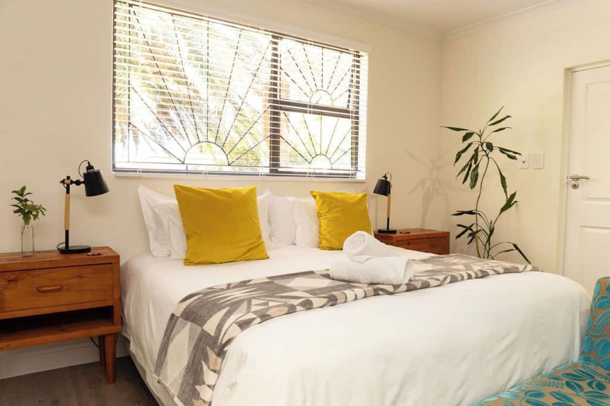 A bedroom with a bed, two bedside tables, a window with blinds, a bedside lamp, a plant, and a door.