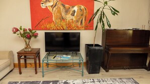 A living room with a television, a coffee table, a sofa, a painting of a lion, and a piano. - Staymore Guesthouse (Stellenbosch)