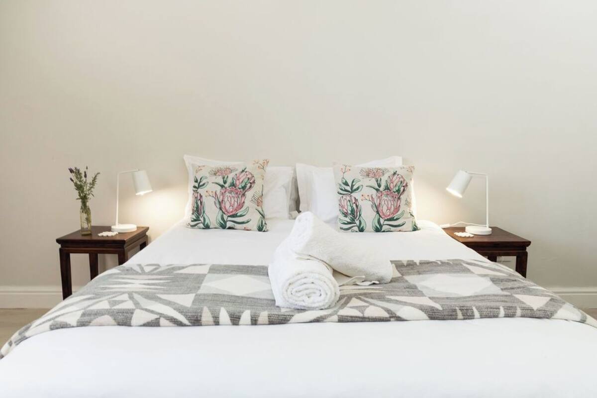 A neatly made bed with floral pillows, a rolled towel, and a geometric-patterned blanket. There are two bedside tables with lamps and a vase of flowers on one.