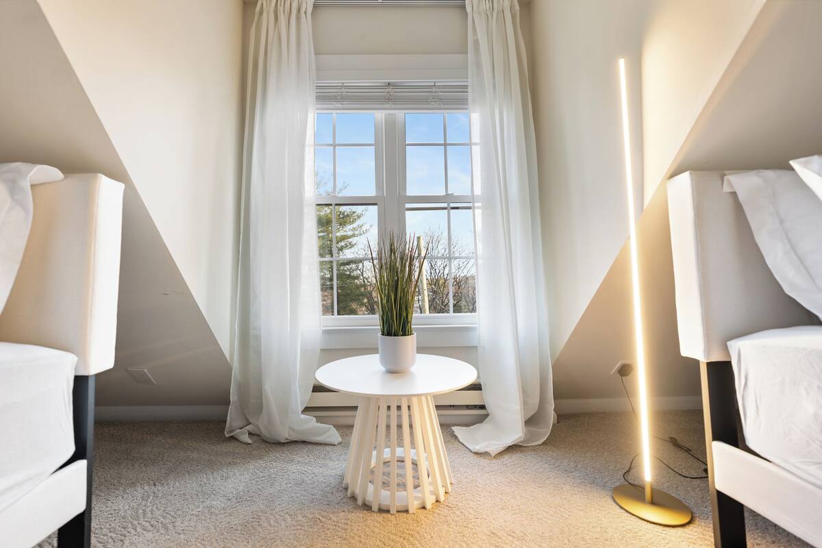 Une chambre moderne avec deux lits blancs, une table ronde blanche ornée d’une plante en pot et un lampadaire.
