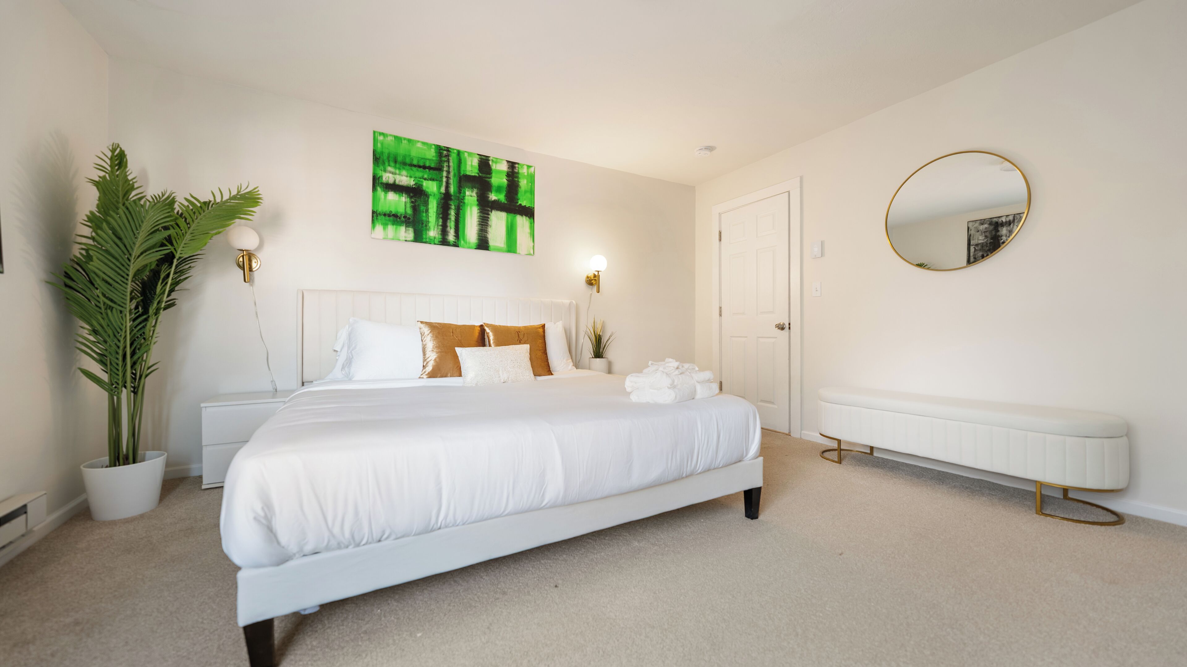 A bedroom with a large bed, a potted plant, a wall-mounted artwork, a bedside table, a nightstand, a radiator, and a round mirror.
