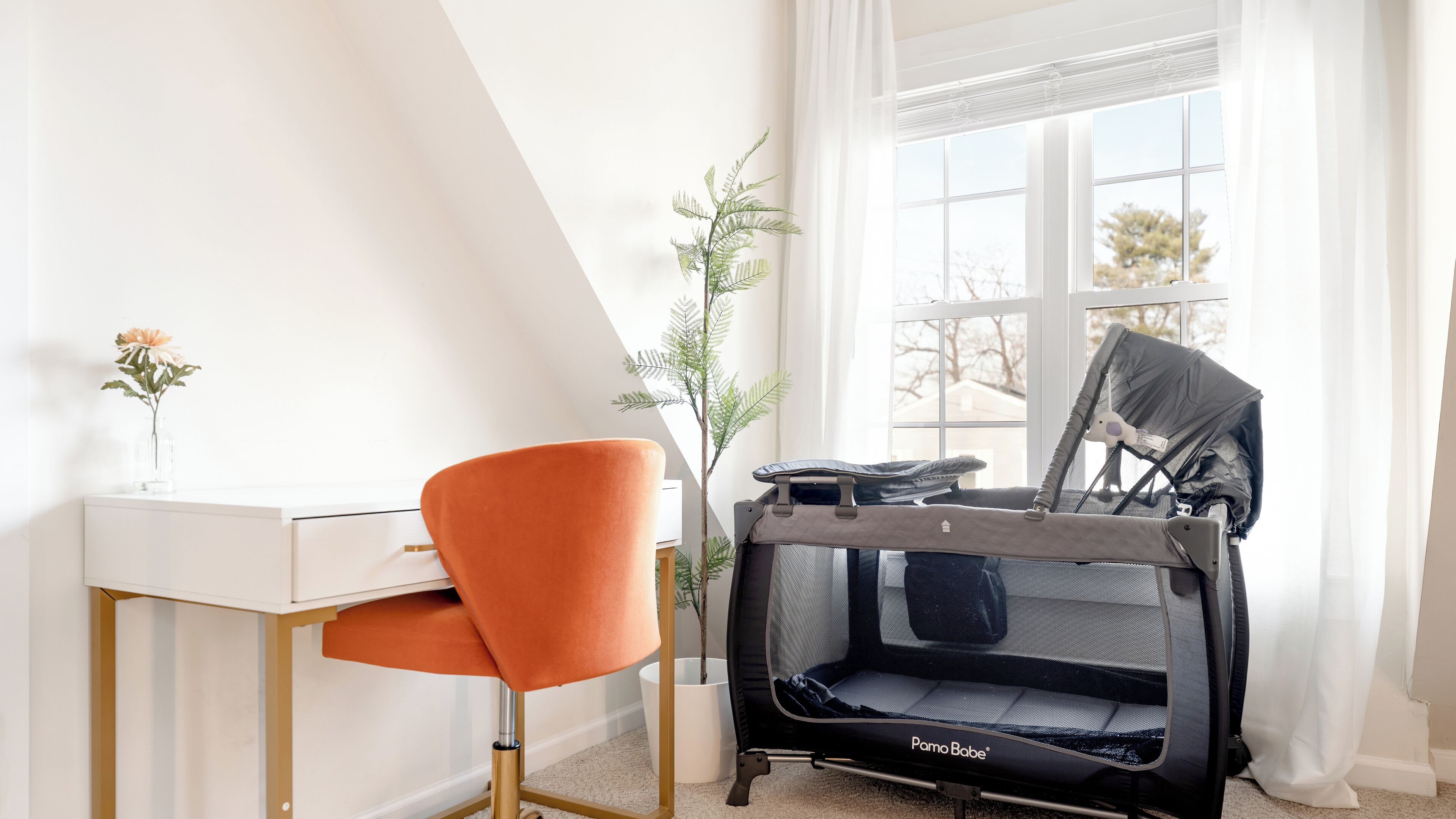 Ein Zimmer mit einem weißen Schreibtisch, einem orangefarbenen Sessel, einem Pama Bebe Kinderwagen, einer Topfpflanze und einem Fenster mit Vorhängen.