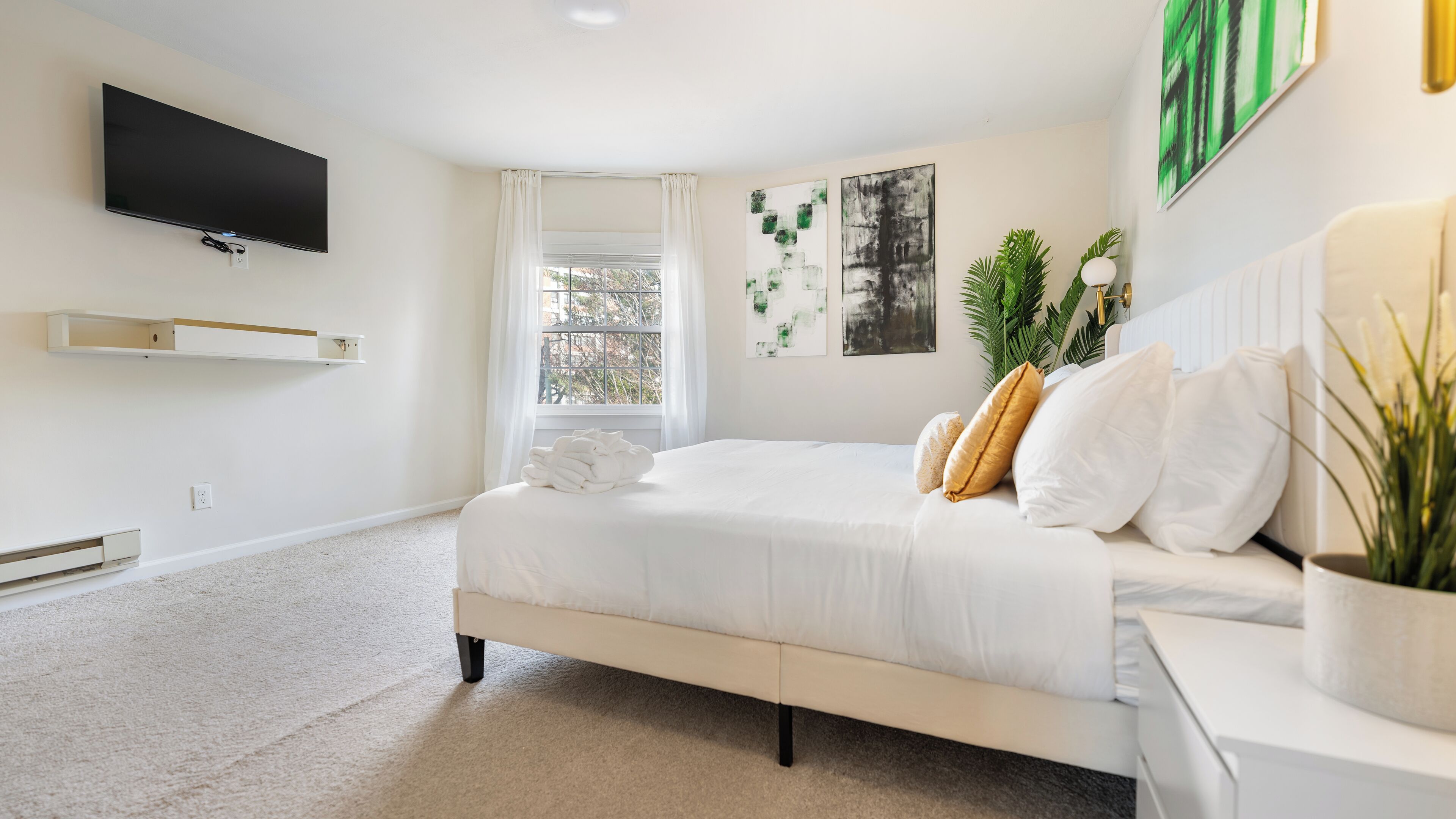 Une chambre à coucher avec un lit, un téléviseur fixé au mur, une fenêtre avec des rideaux et une plante sur la table de chevet.