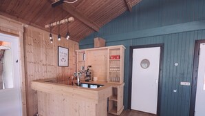 Uma área de bar em madeira, com uma máquina de café, prateleiras com copos e uma porta com um vitral circular.