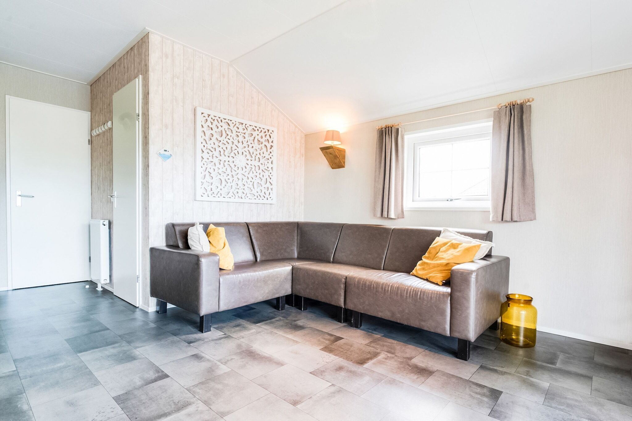 A modern living room with a grey sectional sofa, a decorative wall panel, a floor lamp, and a large window with curtains.