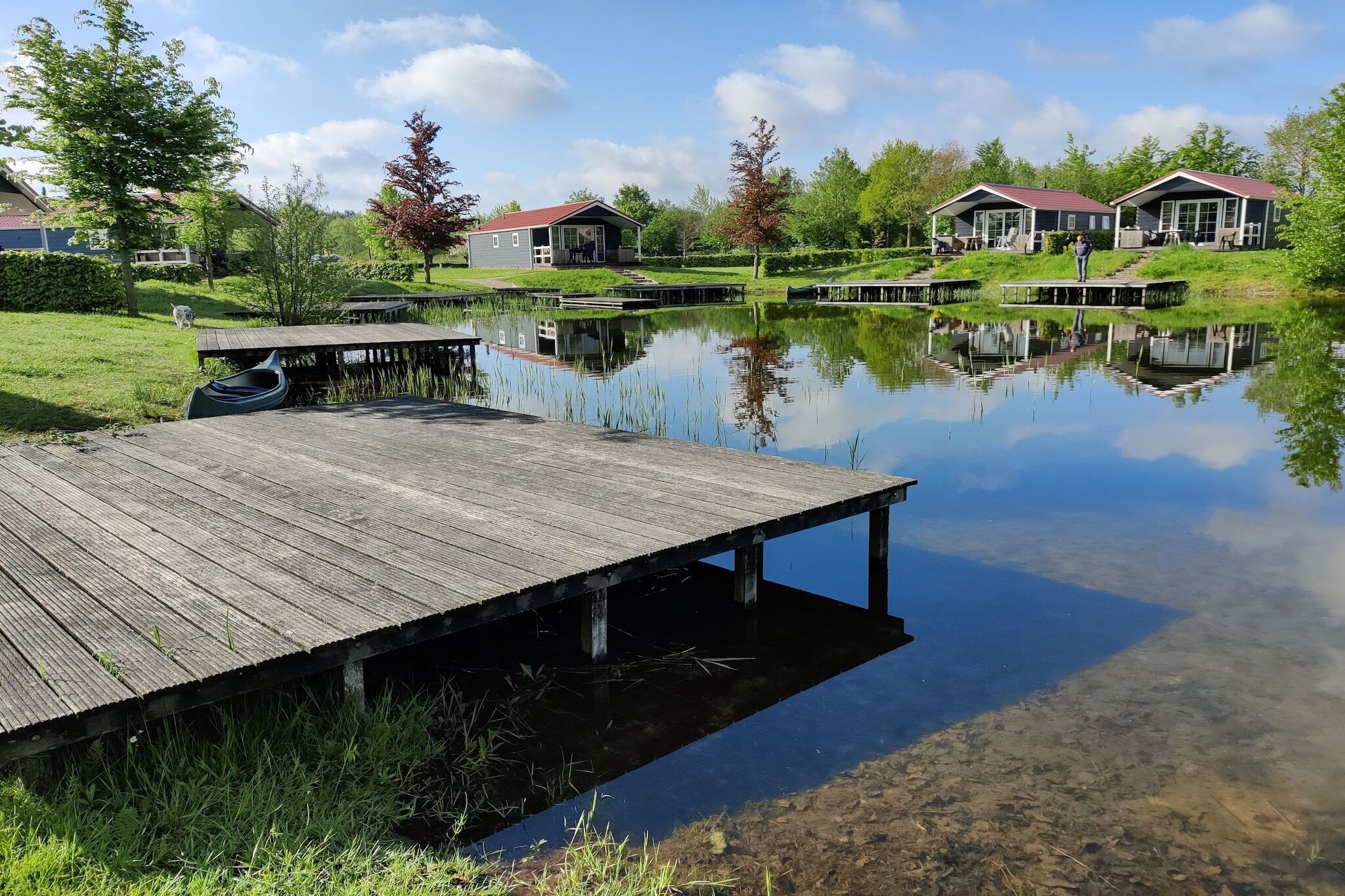 Een houten steiger die zich uitstrekt over een rustig meer, omgeven door weelderige begroeiing en twee huizen met rode daken.