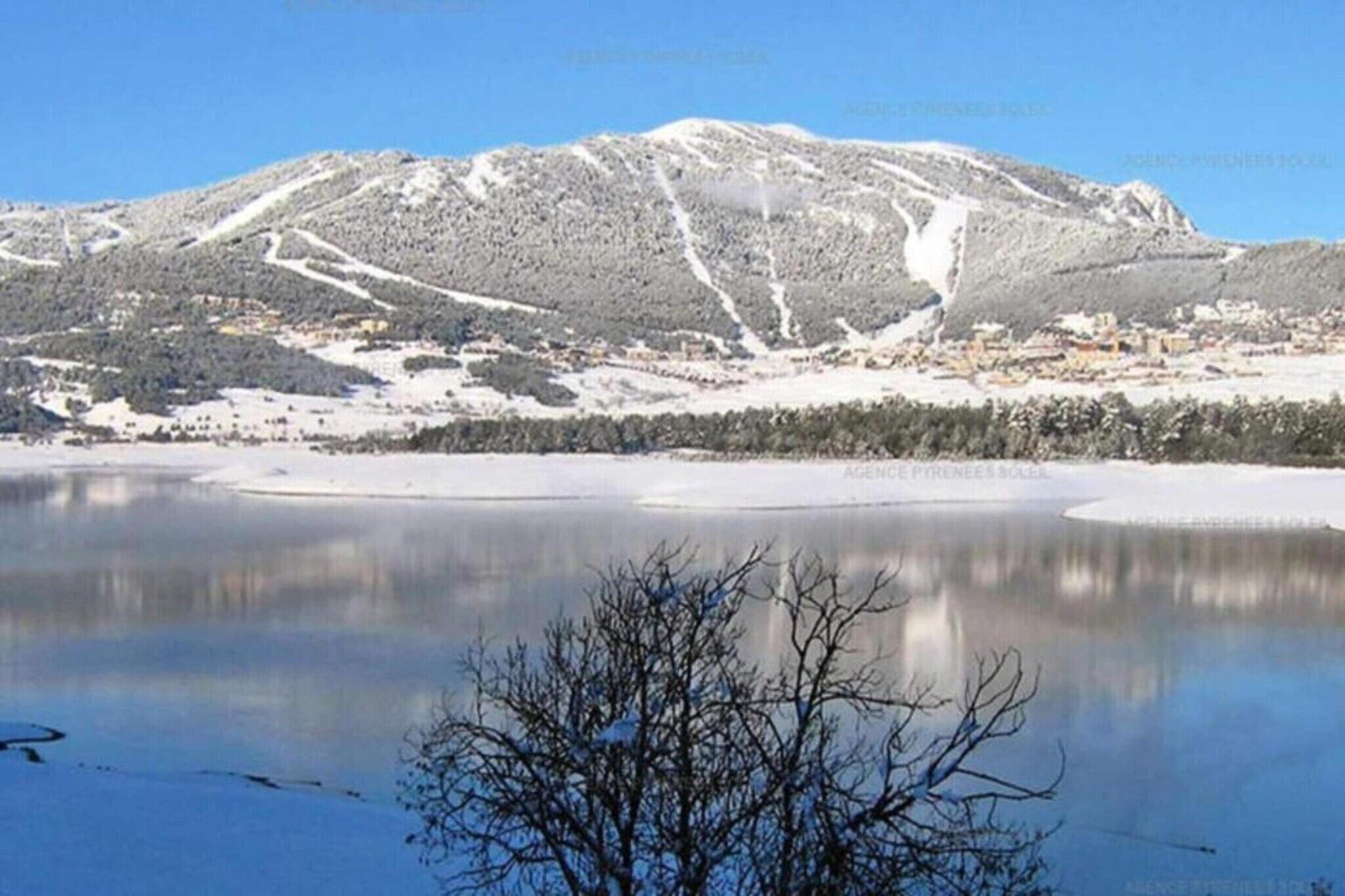 A snow-covered mountain with ski trails and a lake in the foreground.