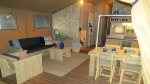 A rustic cabin interior with a wooden floor, a sofa, a dining table with chairs, a small kitchen area, and a clock on the wall.
