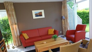 Un salon avec un canapé rouge, une table basse en bois, des fauteuils en osier et une grande fenêtre donnant sur un paysage verdoyant.