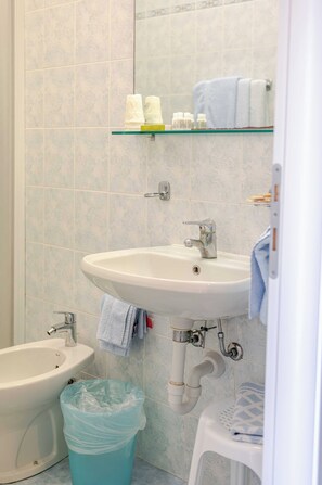 A bathroom with a white sink, a bidet, a glass shelf with toiletries, and a trash bin.
