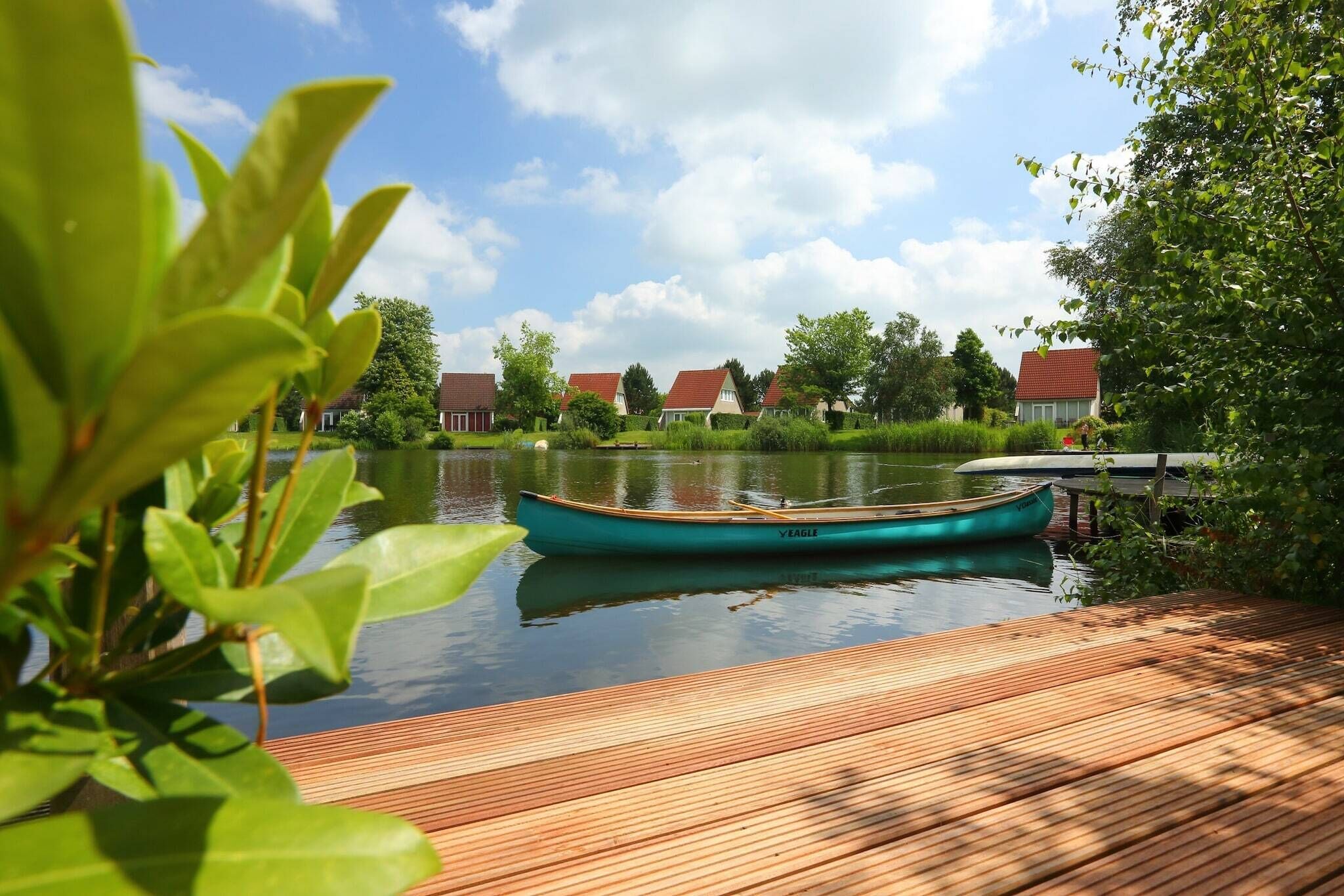 Ein hölzerner Steg mit einem dort angelegtem Ruderboot namens "VEAGLE" an einem ruhigen See, umgeben von Grünflächen und Häusern mit roten Dächern.