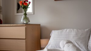 Una cama bien hecha con sábanas blancas y una toalla blanca enrollada, un aparador de madera y un jarrón con flores rojas sobre una mesa.