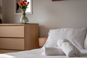 A neatly made bed with white linens and a rolled white towel, a wooden dresser, and a vase with red flowers on a table. - James Robb Avenue (No 4) (St Andrews)