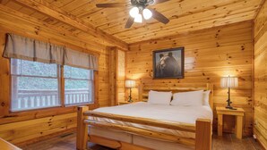 A wooden cabin bedroom with a bed, two bedside lamps, a ceiling fan, and a window with blinds. - Sleeps 28 | Indoor Pool, Theater, Game Room, Hot Tub, Views | Marco Polo Lodge (Sevierville)