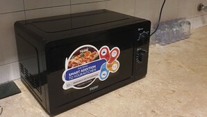 Un horno tostador Haier Solo Series negro sobre una encimera de cocina con salpicadero de azulejos y un enchufe eléctrico al fondo.